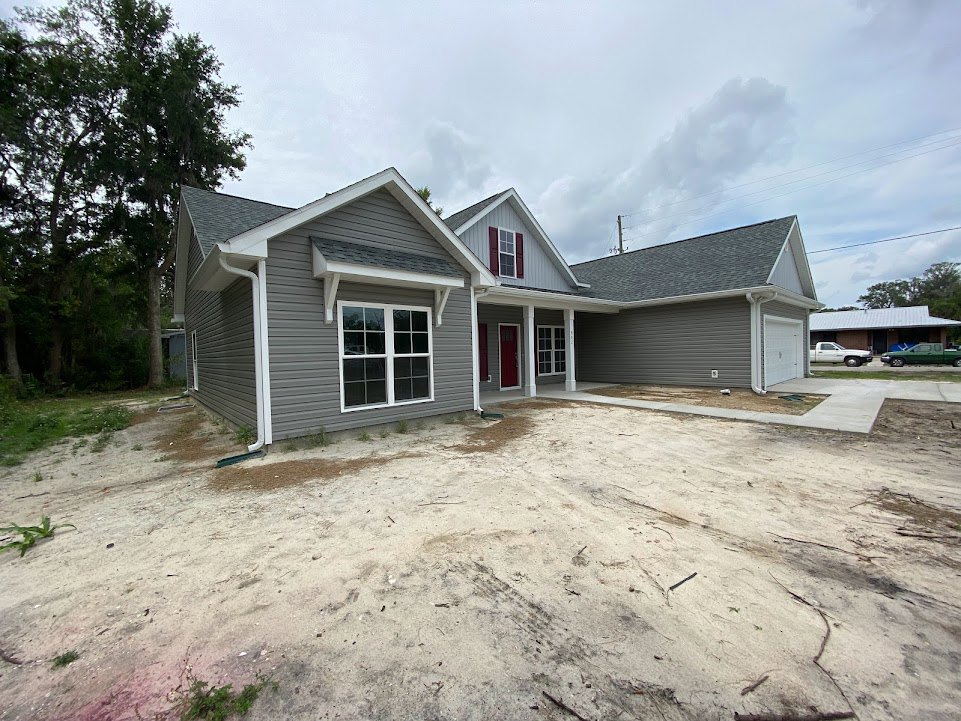 Grey siding house with white trim, red front door, white-framed windows, concrete driveway, dirt landscaping, mature trees in background, cloudy sky overhead