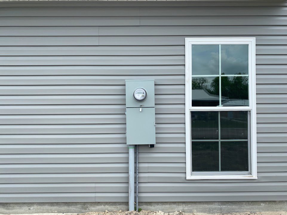 Grey electrical meter box mounted beside a white-framed window on exterior grey siding of a residential home.