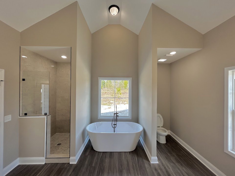 Bathroom with white square tub and metal shower pole, white toilet with seat up, tiled floor, window with white frame and outdoor view, ceiling light fixture, neutral wall tiles.
