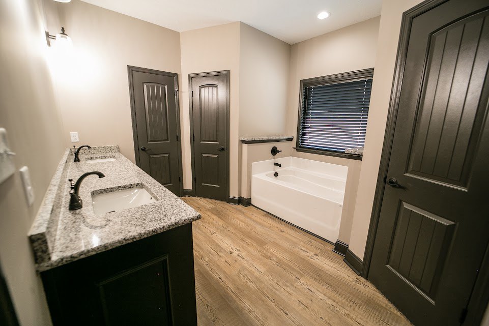 Freestanding white bathtub beside a window with white blinds, modern bathroom sink with chrome faucets, light tile flooring, grey door with black handle, white cabinetry and