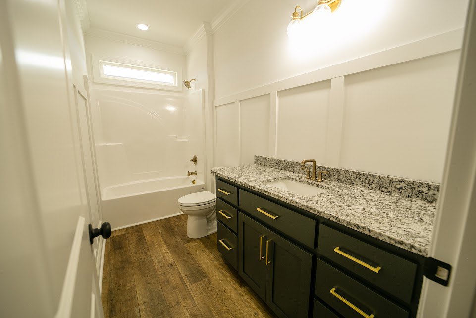 Bathroom with wood flooring, white toilet, white sink set in cabinetry, black door knob on white door, and drawers adjacent to toilet.