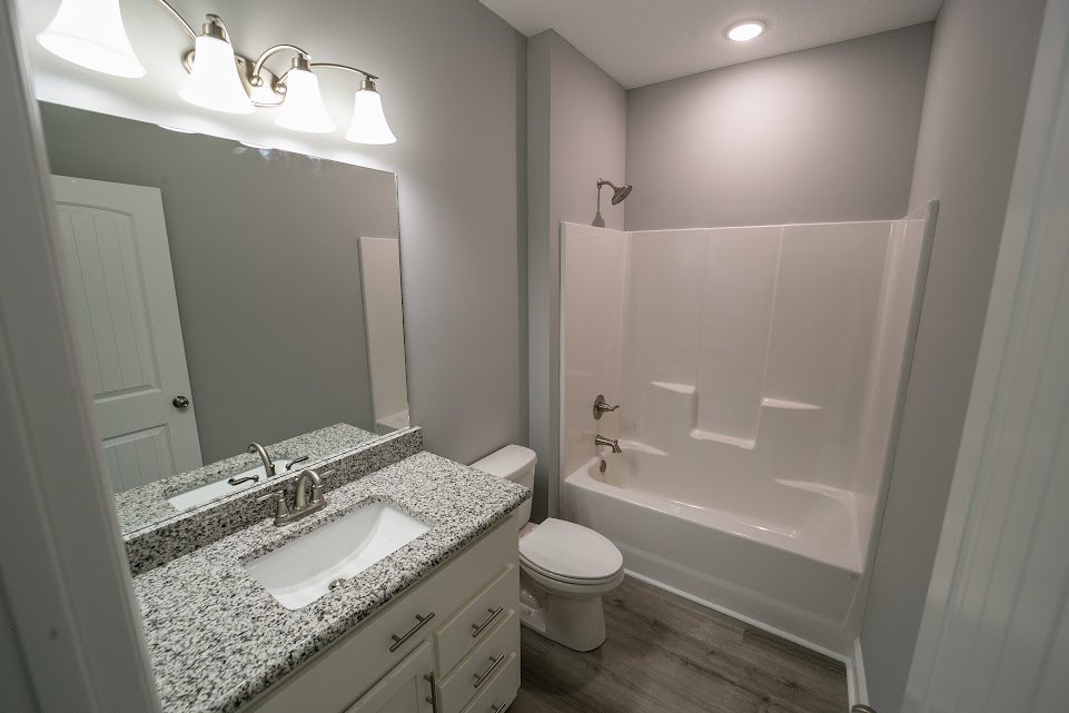 Modern bathroom with white tile walls, a white toilet beside a rectangular bathtub, a white sink with a silver faucet, wall-mounted mirror, and chrome light fixture above the