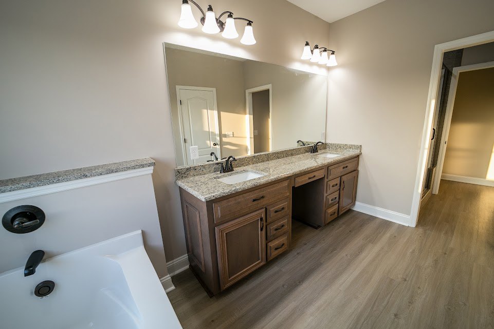Bathroom with a wide mirror above double sinks, white freestanding tub with black handle, row of lights mounted over mirror, tiled floor, cabinetry, and a window.