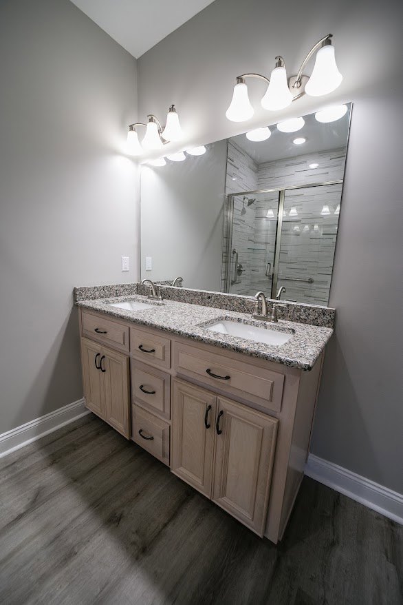 Double vanity bathroom with white sinks, marble countertop, large framed mirror, light gray tile backsplash, wood cabinetry, chrome faucets, and recessed lighting.