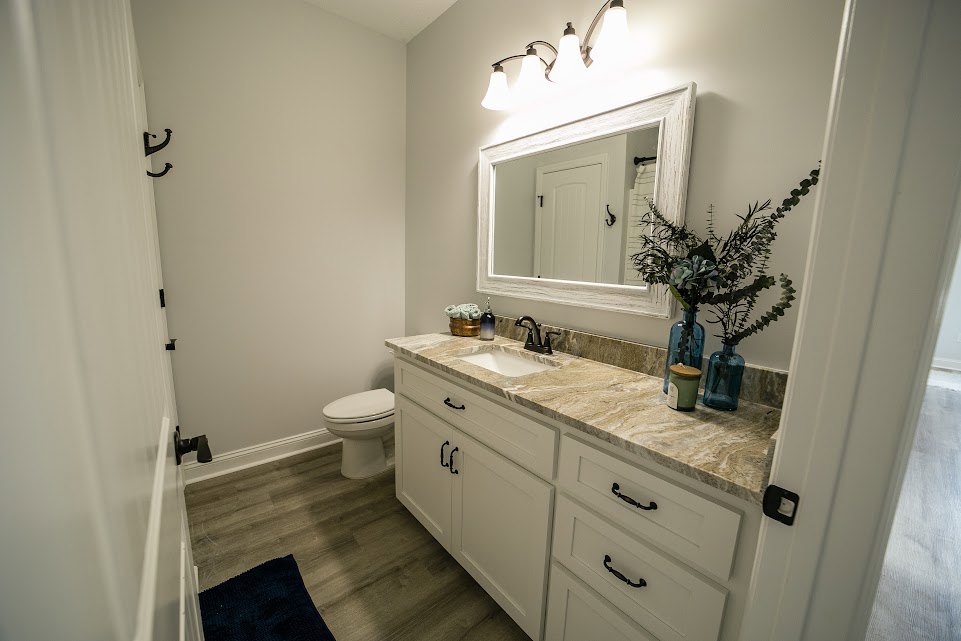 Marble countertop with built-in sink, wall-mounted mirror, blue vases holding greenery, modern light fixture, white toilet, and tiled walls in a contemporary bathroom.