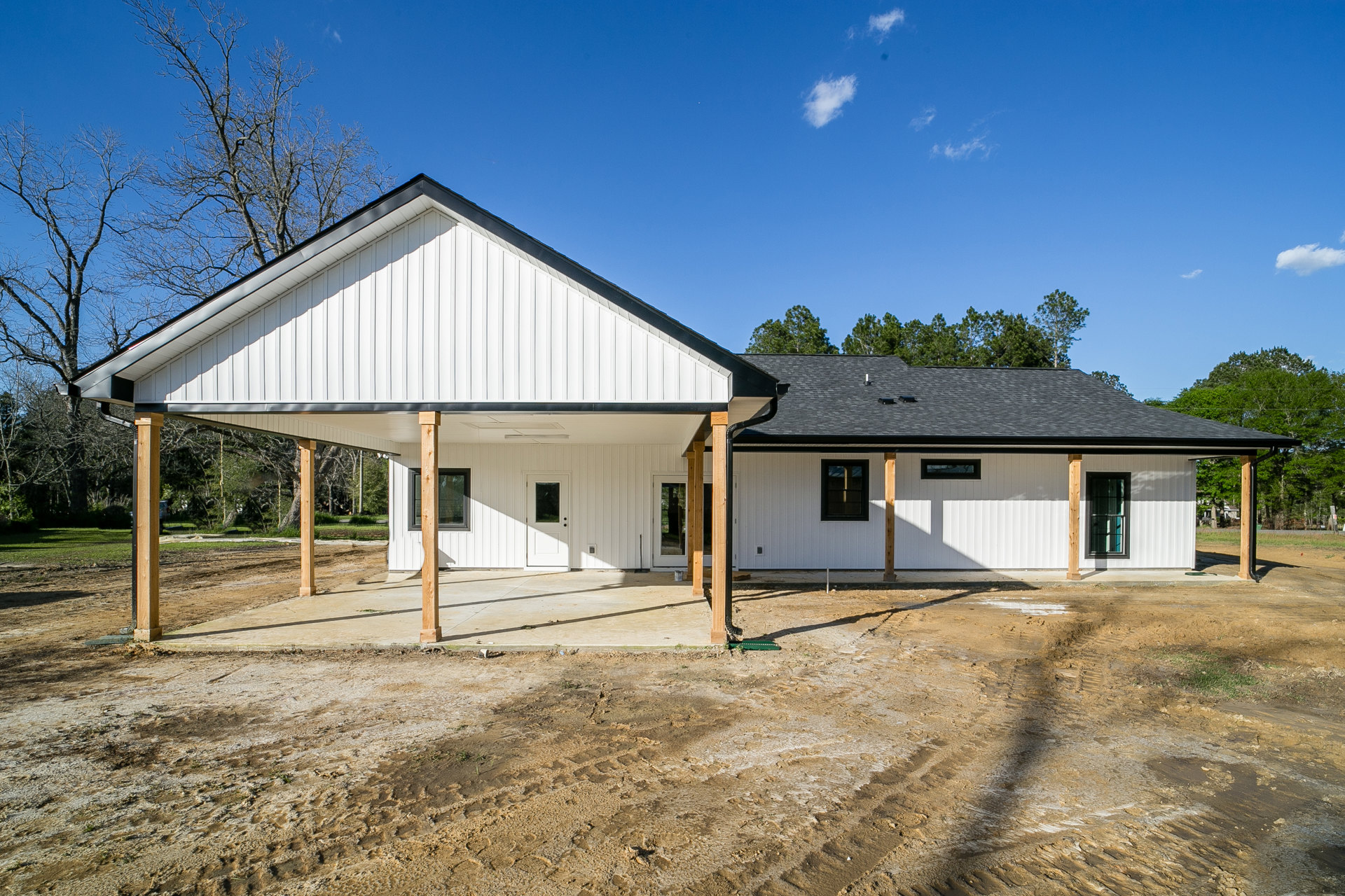 Two-story house under construction with exposed framing, white and black window frames, covered porch, dirt lot in foreground, scattered construction materials, cloudy sky