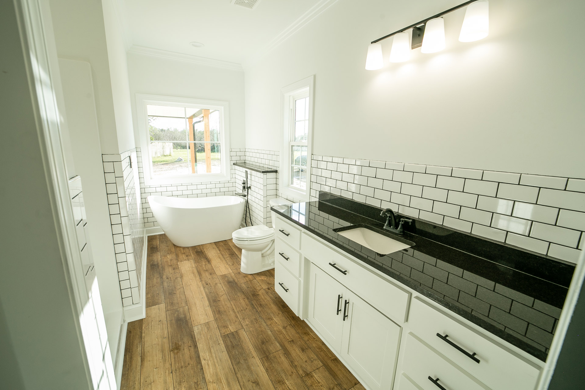 White freestanding bathtub beside black countertop vanity, closed toilet, row of wall-mounted lights, window with white frame, light tile flooring and cabinetry