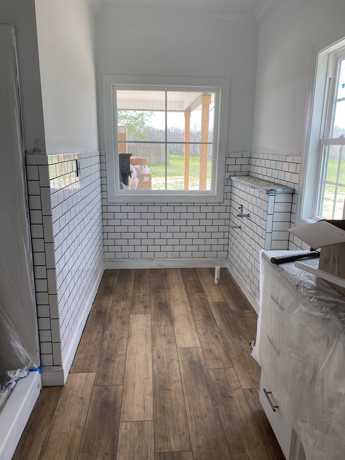 Sunlit room featuring a large window overlooking a green yard, wood flooring, white tile walls, a table draped with a white sheet, wood shelving, and stacked moving boxes.