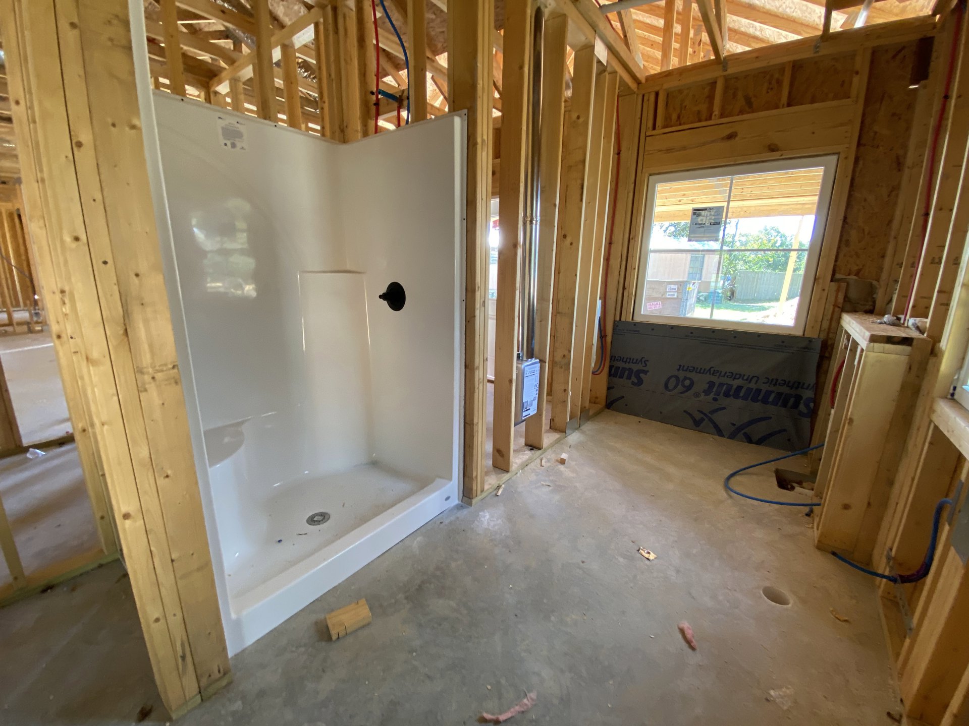 Bathroom under construction with exposed wooden ceiling beams, unfinished plaster walls, white shower unit, and window displaying a sign.
