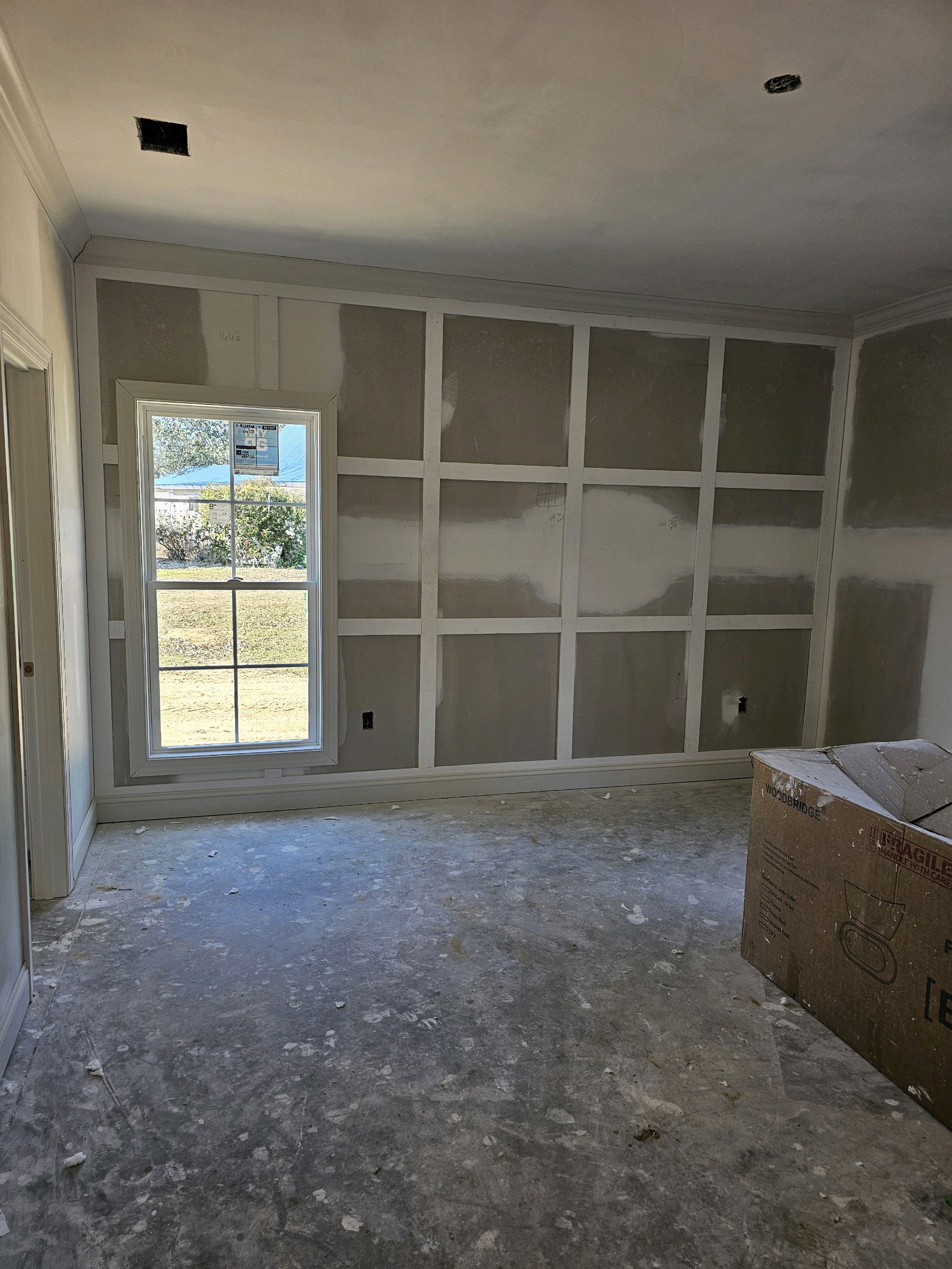 White plaster walls and ceiling, large window with sign, cardboard box on light-colored floor, sparse furnishings in empty room