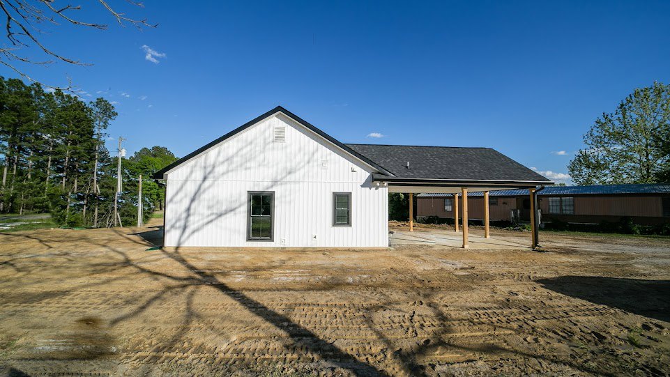 White siding house with black shingle roof, surrounded by dirt ground with tire tracks, dense forest of green-leaved trees in background, cloudy sky overhead.