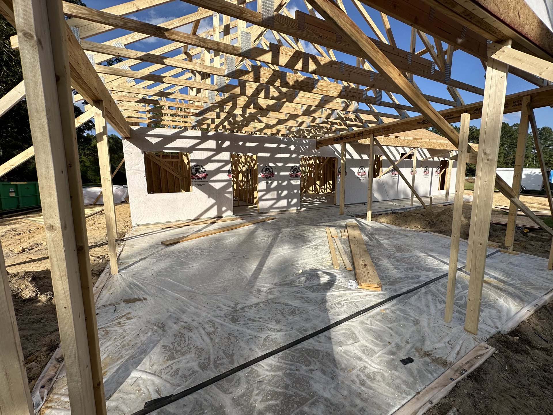 Wood-framed house under construction with exposed beams, concrete floor, partially completed roof, and green container with yellow label on site