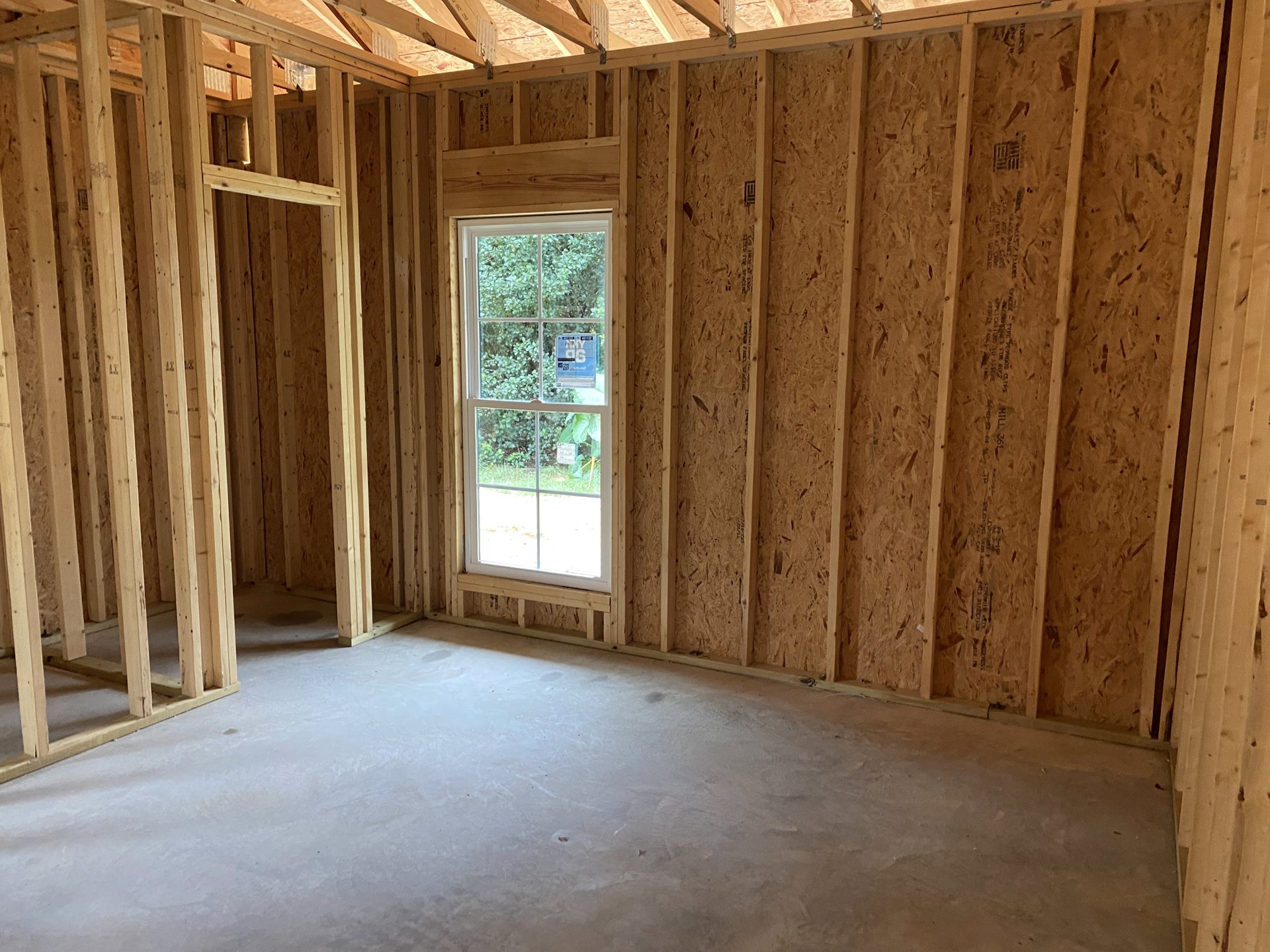 Room with exposed wooden ceiling beams, metal brackets, concrete floor, large window with blue and white sign, unfinished walls.