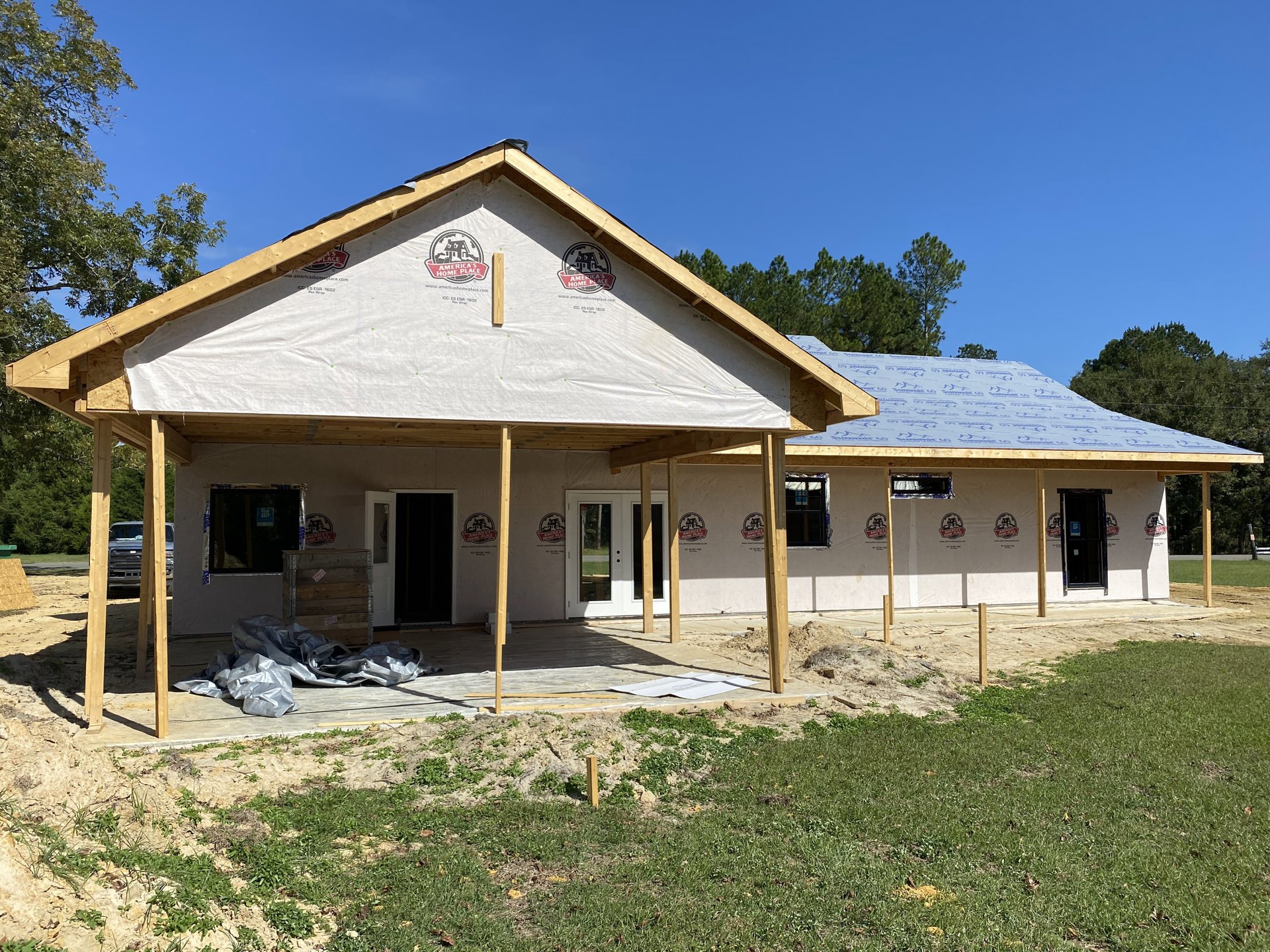 Two-story house under construction with exposed framing, covered porch, and green lawn bordered by trees under a partly cloudy sky