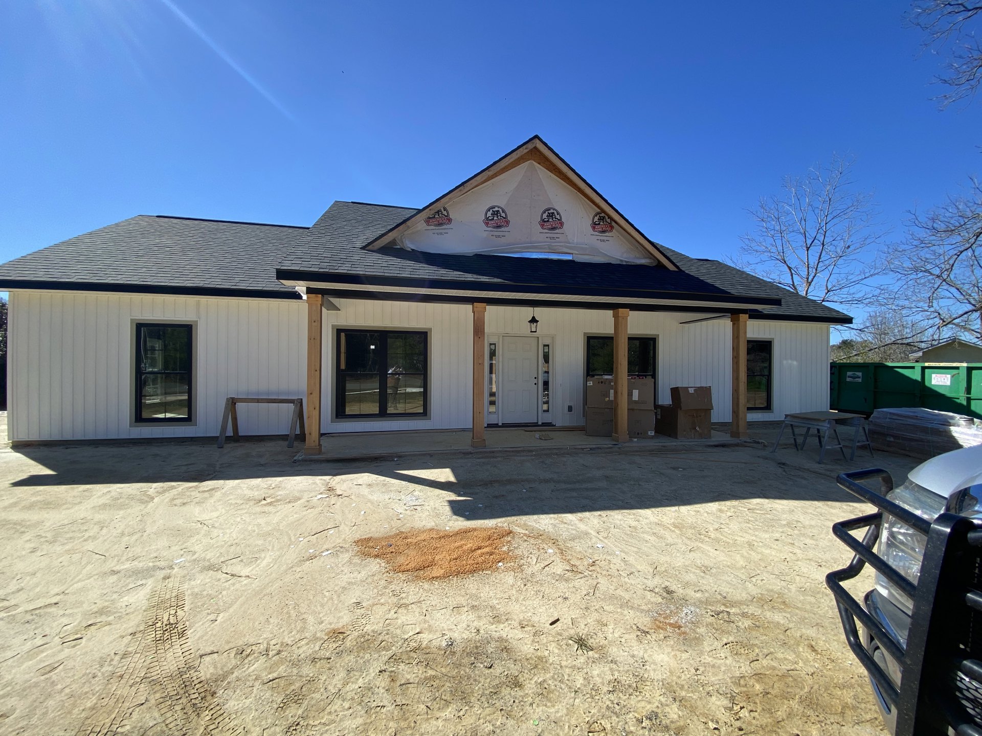 White siding house with black framed windows, white tarp partially covering roof, lawn chair on grassy front yard, pile of dirt near driveway, clear sky overhead.