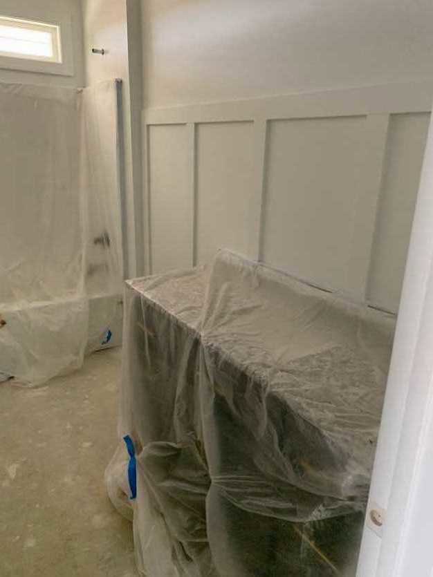 Dining table draped in clear plastic sheeting, white paneled wall and window with illuminated light fixture in background, partially visible blurry face, grey surface in