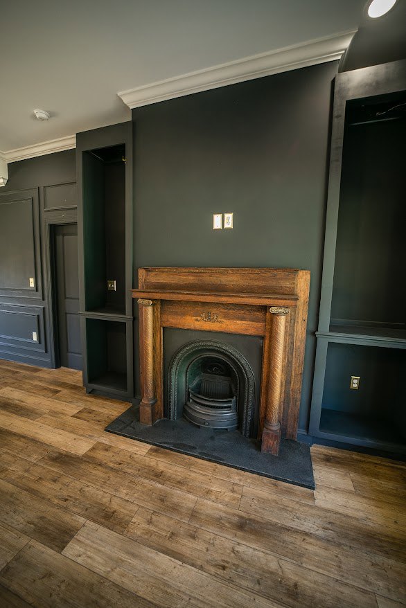 Wood-burning fireplace with a wooden mantle set against a neutral wall, hardwood flooring, and fire screen in a cozy living room.