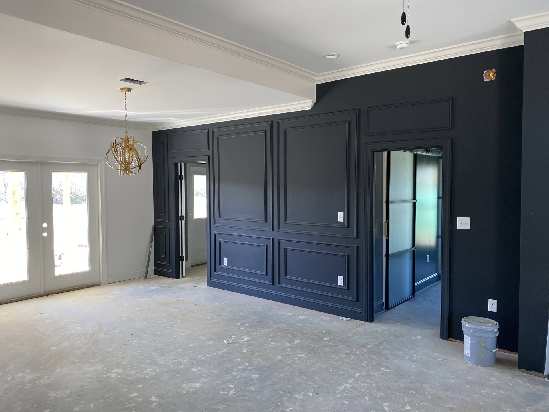 Black-walled room with a gold chandelier hanging from a chain, glass-paneled door, concrete floor, and white paneled accent wall.