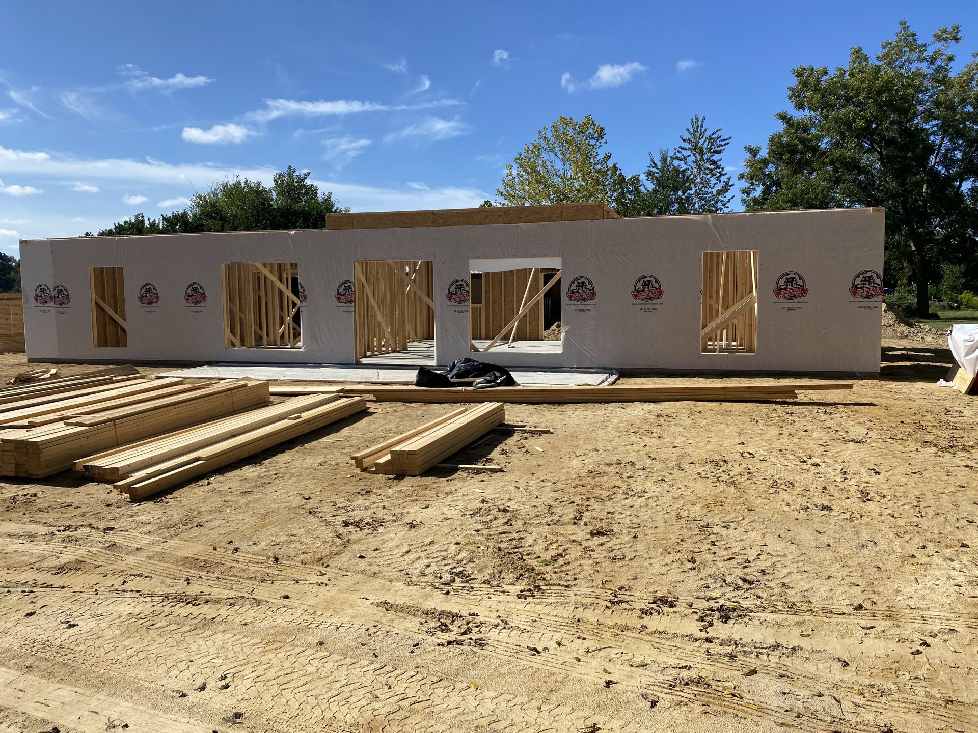 Wood-framed custom home under construction, exposed beams and studs, stacks of lumber and black tarp on dirt lot, cloudy sky and trees in background