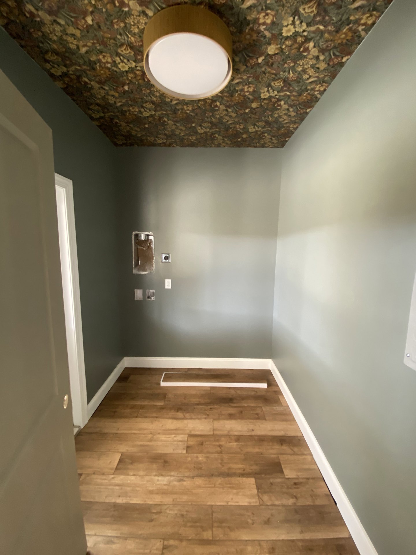 Ceiling-mounted circular light fixture on a floral-patterned ceiling, wood flooring with white trim, partial view of a metal pole with a brown glove, white wall with a black spot