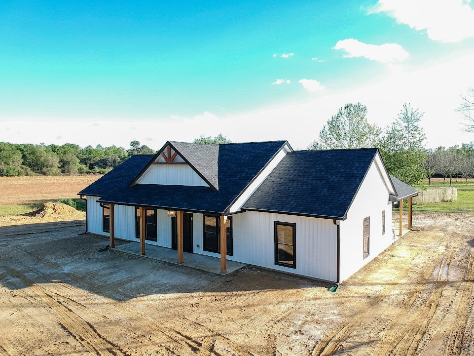 White house with black roof under construction, dirt driveway in foreground, blue sky with clouds overhead, window reflecting surroundings, close-up of roof shingles visible.