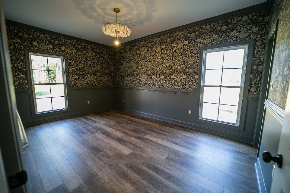 Chandelier with white flower-shaped bulbs hanging from plaster ceiling, wood flooring, large windows with white frames overlooking trees, decorative molding along walls