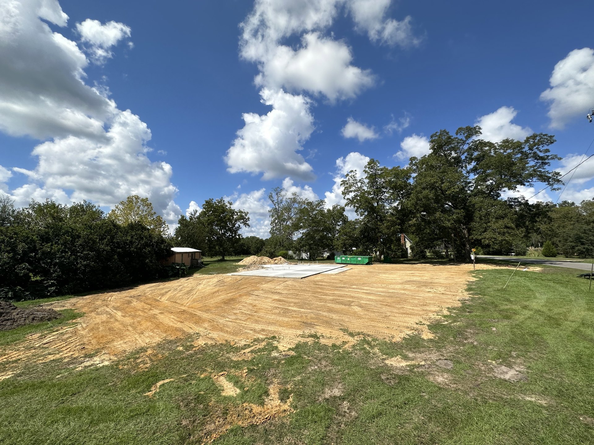 Dirt field bordered by trees under a blue sky with scattered clouds, small shed partially visible among woods, green stake in grassy area near house