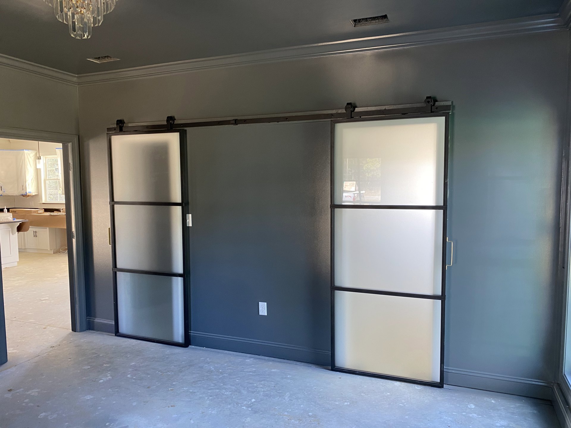 Sliding glass doors with black frames set in a white wall, crystal chandelier overhead, grey accent wall, light wood flooring, and partial view of a refrigerator.
