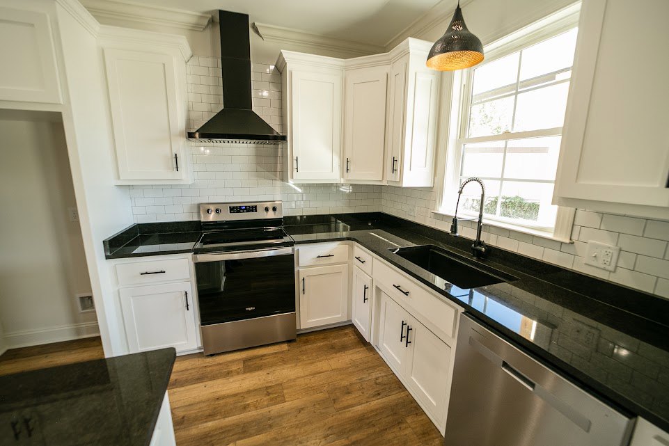 White cabinetry and black countertops in a modern kitchen, stainless steel stove and dishwasher, black and gold pendant light fixture, undermount sink beneath ceiling light, lamp