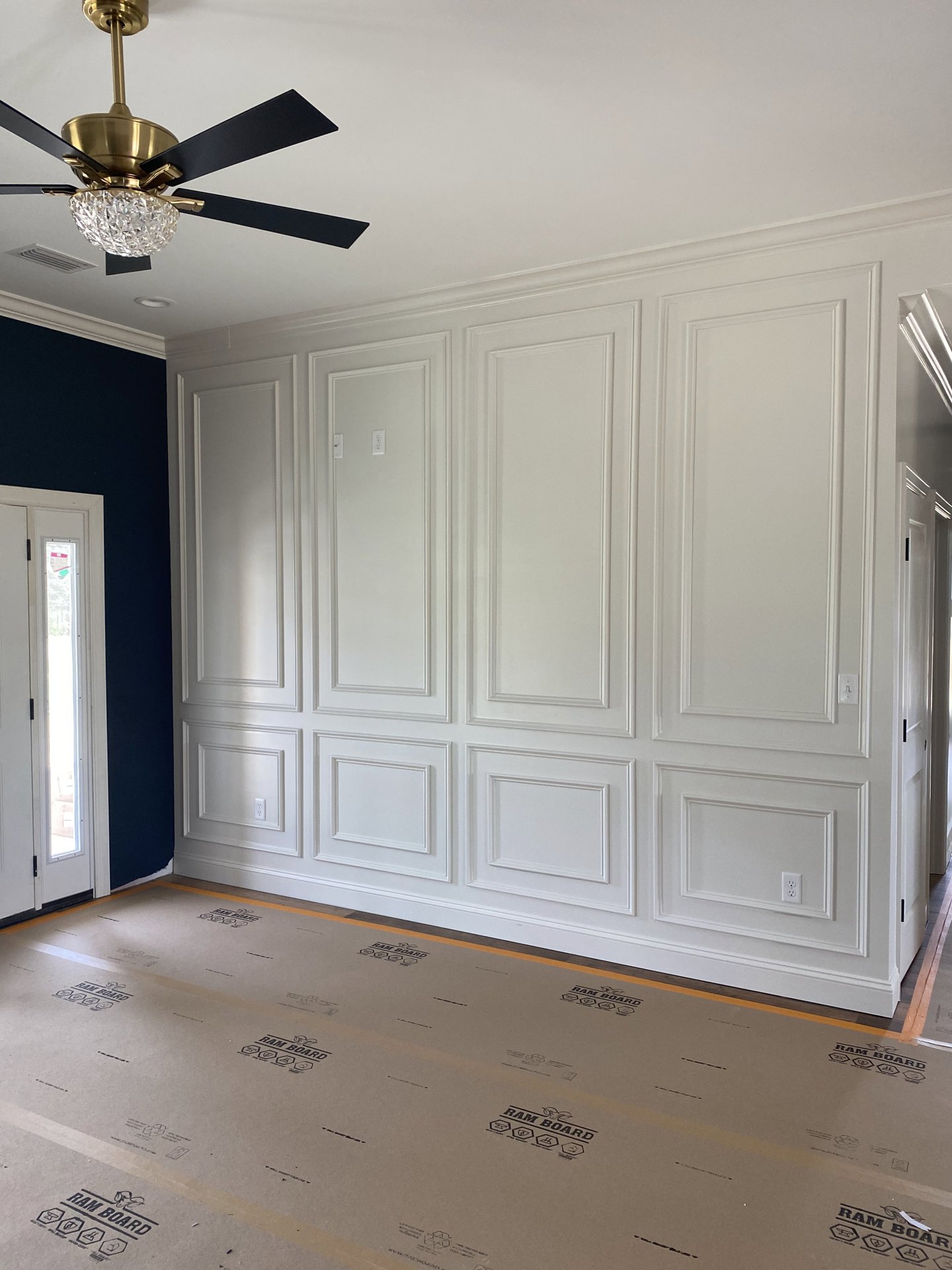 Ceiling fan with black blades and crystal ball fixture, white paneled walls, cardboard floor with black logo, two white doors with light shining through one