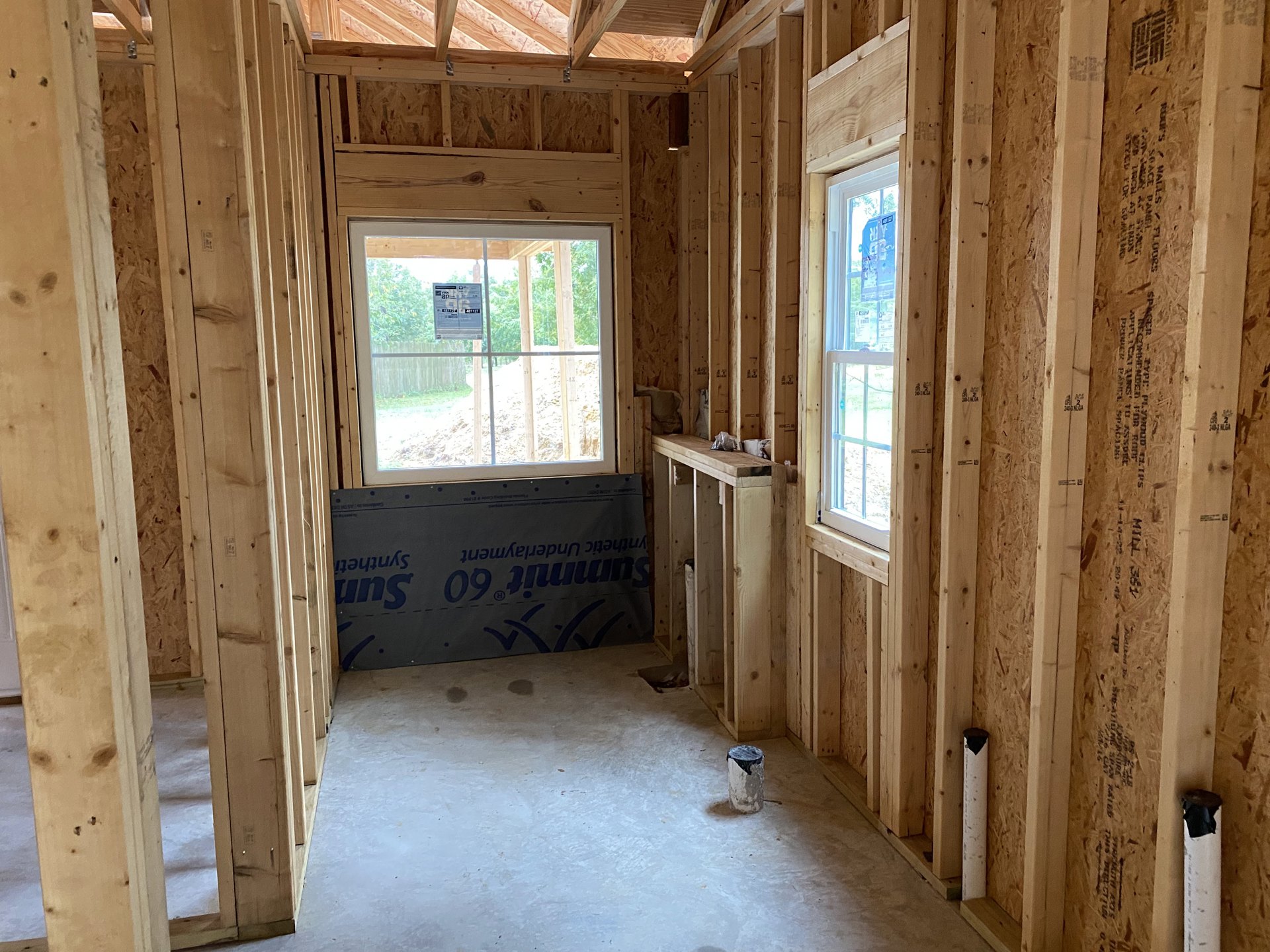Unfinished room with exposed wooden beams, concrete floor, and window displaying a grey sign with blue writing