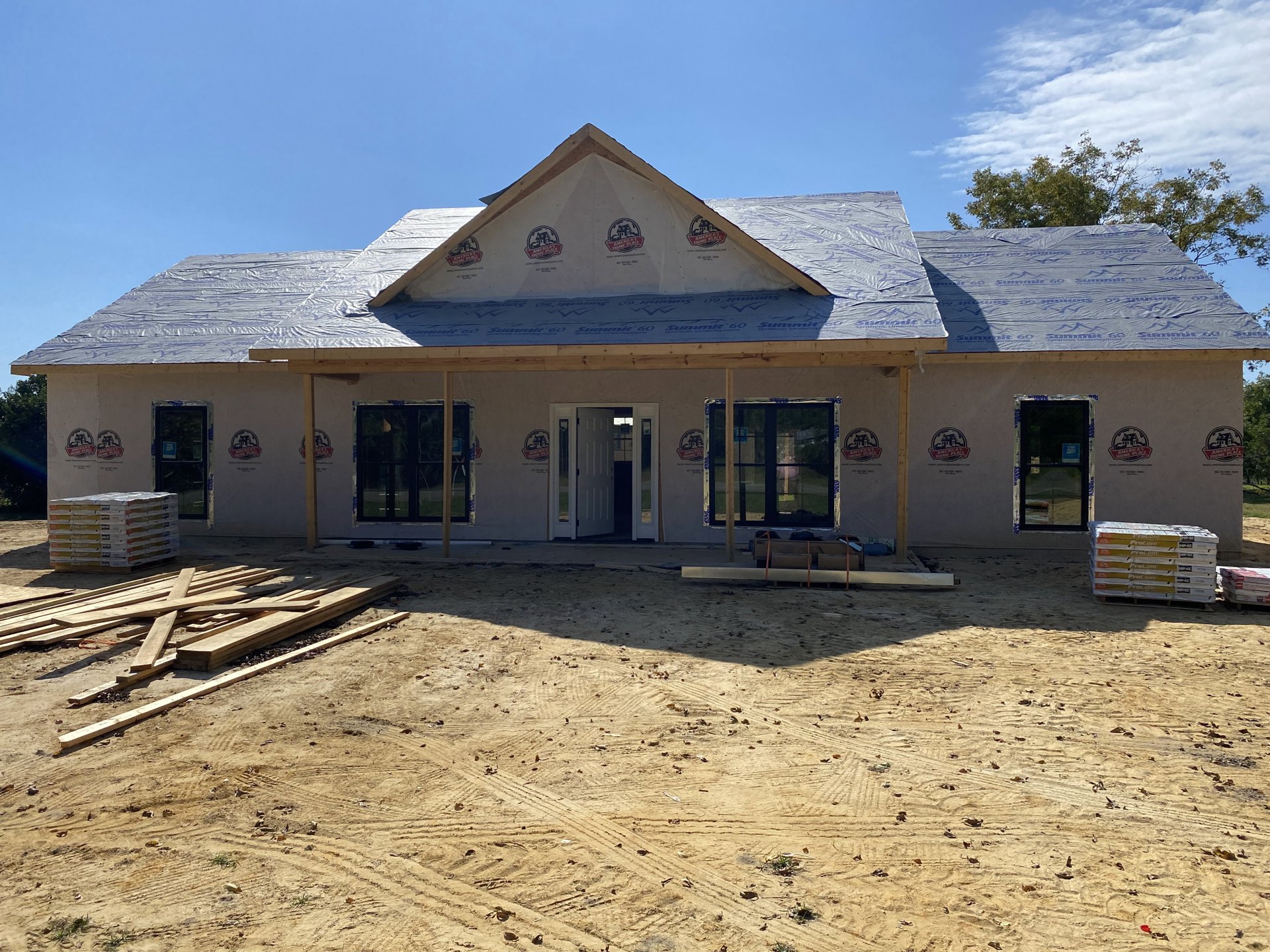 Partially built house with exposed framing, dirt yard with tire tracks, stack of white and yellow construction boxes, window being installed with sign, exterior door, cloudy sky