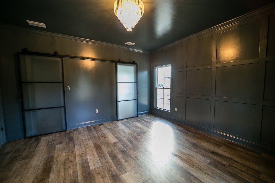 Spacious room featuring wide-plank wood flooring, a modern chandelier, white-framed windows, and a glass door with a black frame.