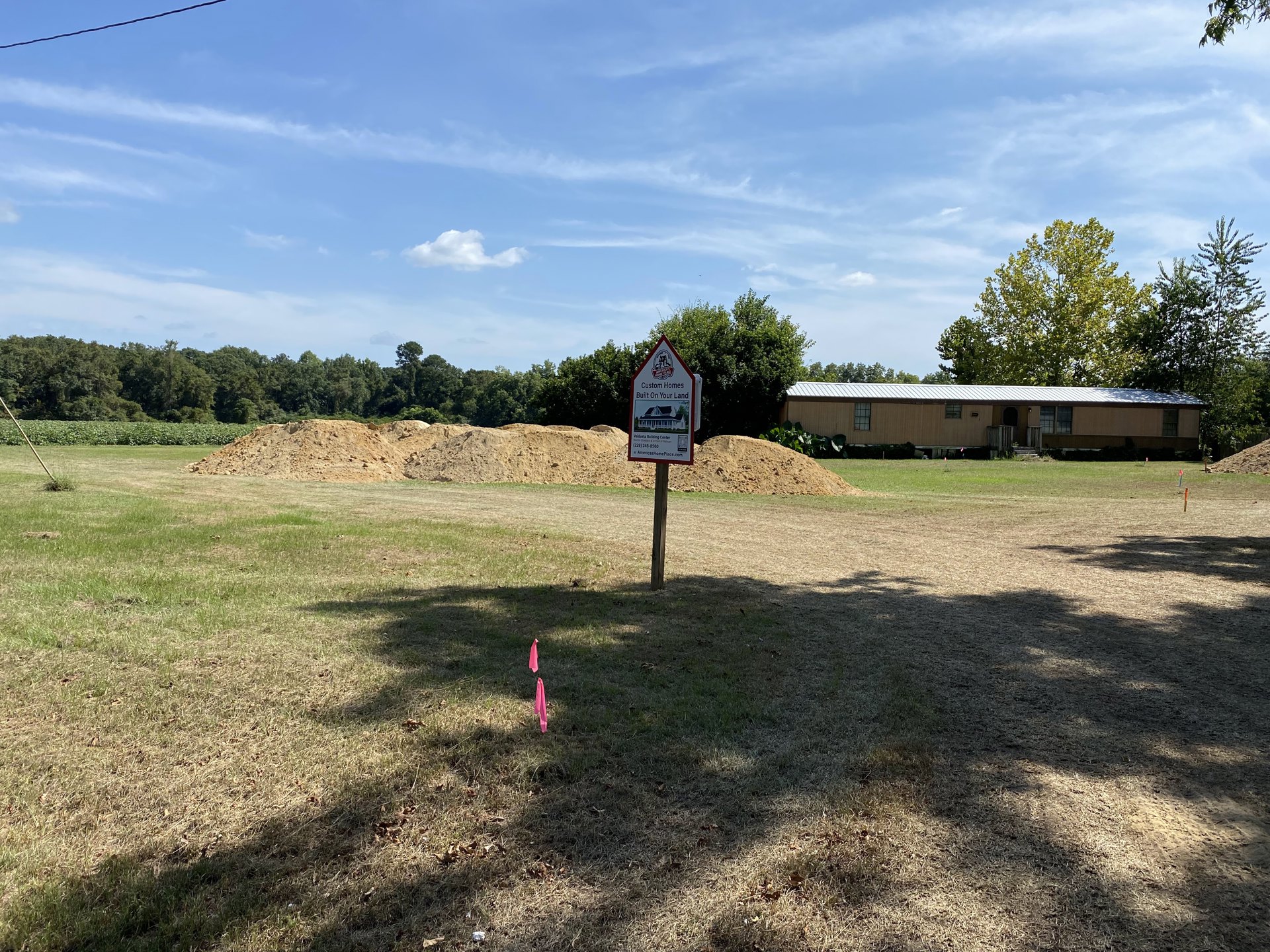 White sign with house illustration mounted on wooden post in grassy field, two-story home with porch and stairs in background, red and white triangular roof, mature trees, partly