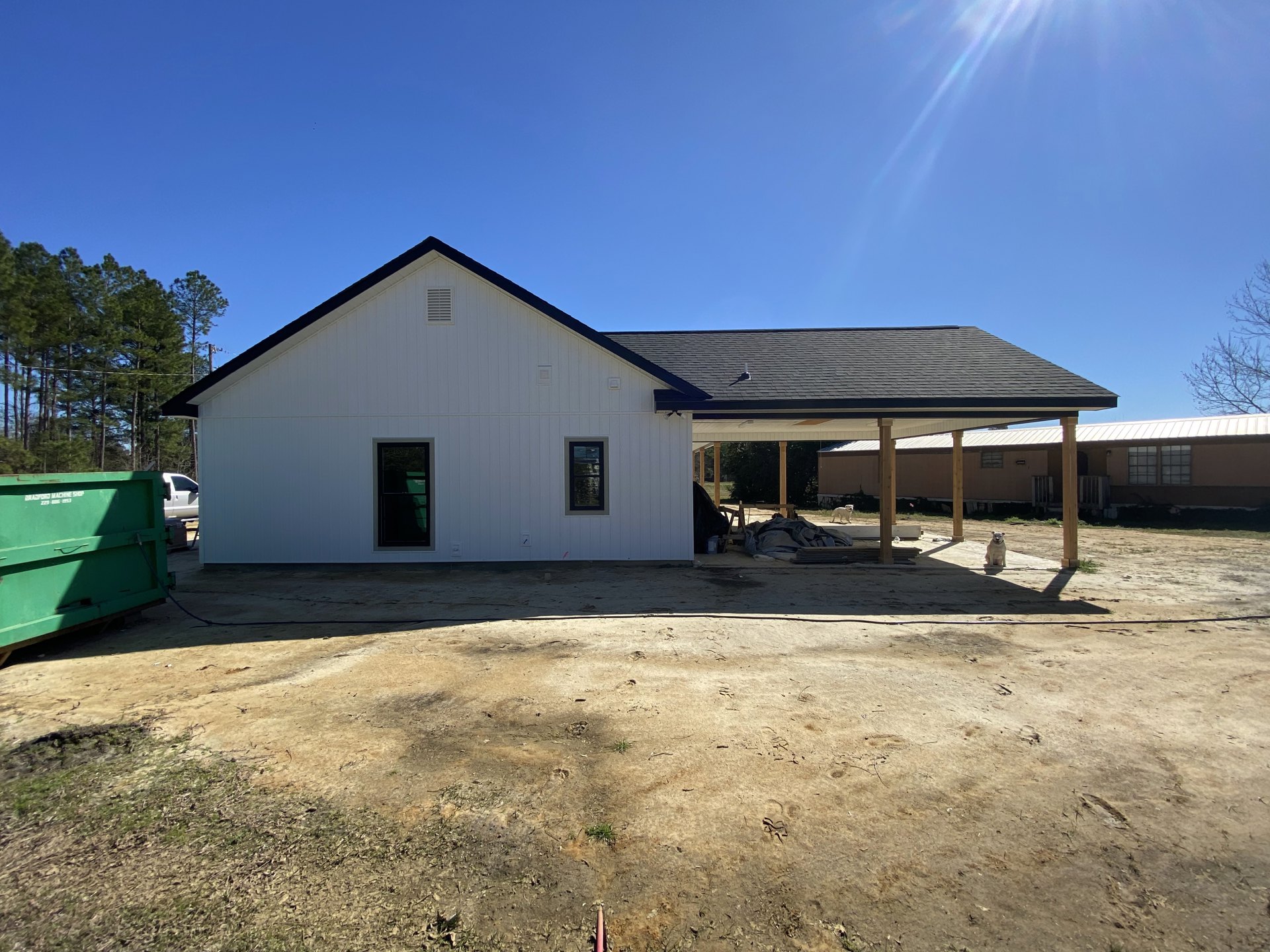 White house with black roof under construction, covered patio supported by columns, green dumpster and hose on dirt area in foreground, windows reflecting nearby trees, cloudy sky