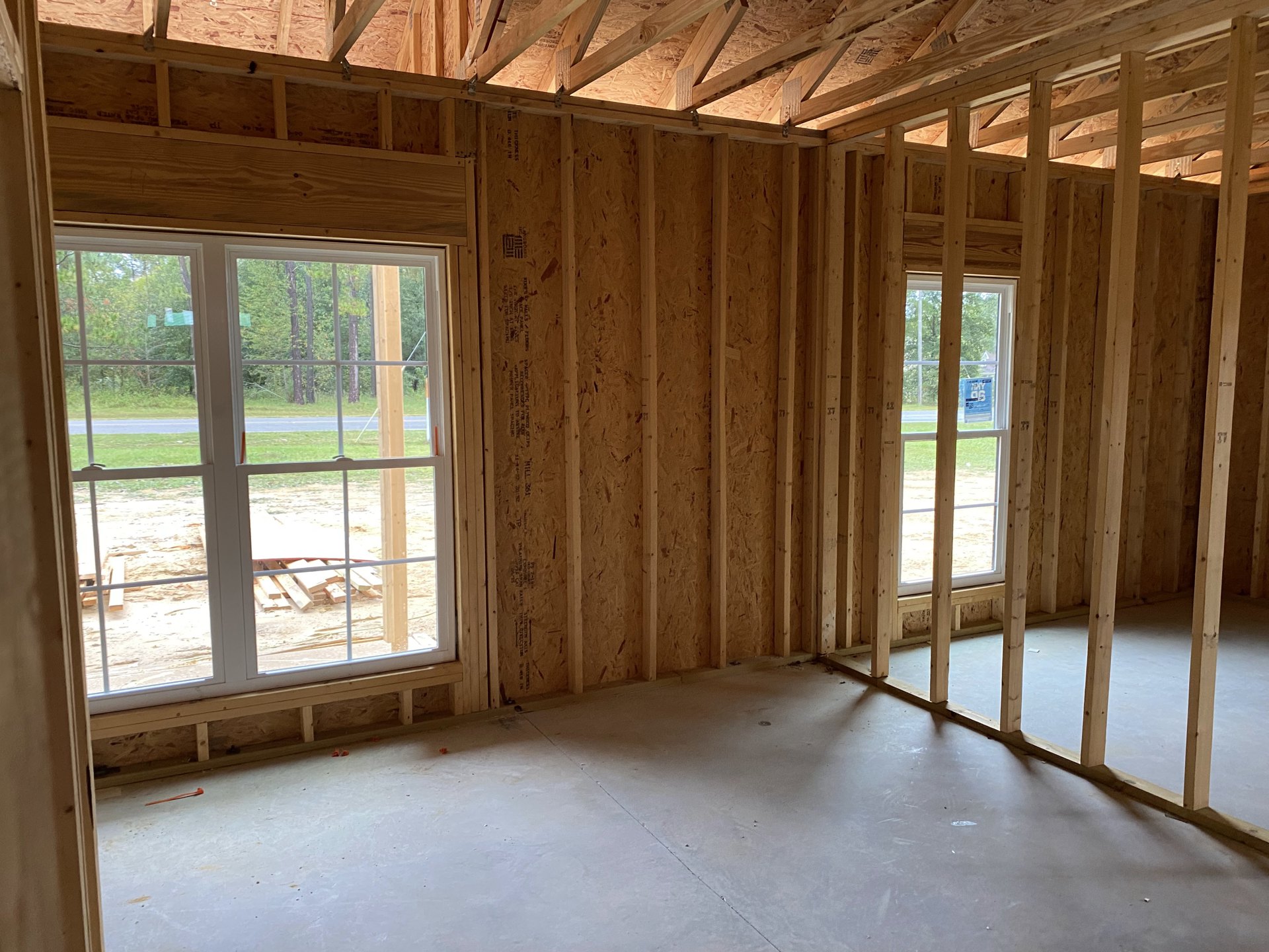 Spacious room featuring large windows overlooking a yard with trees, concrete floor, exposed wooden ceiling beams, and a pile of lumber on a table.