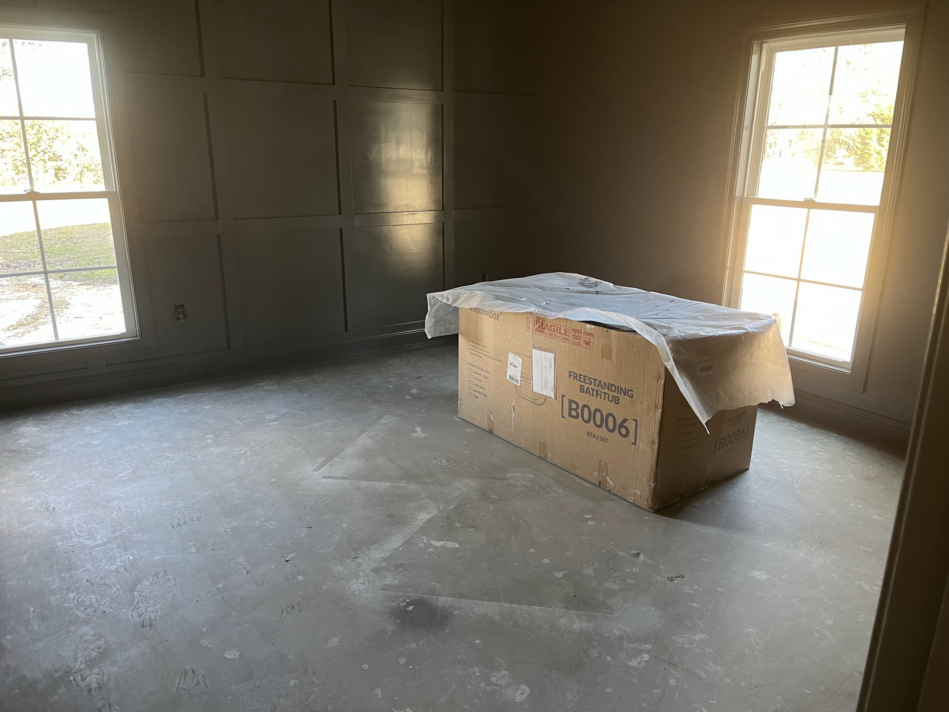 Large cardboard box with plastic covering placed on light-colored floor in empty room, white-framed windows on walls, natural light streaming in.