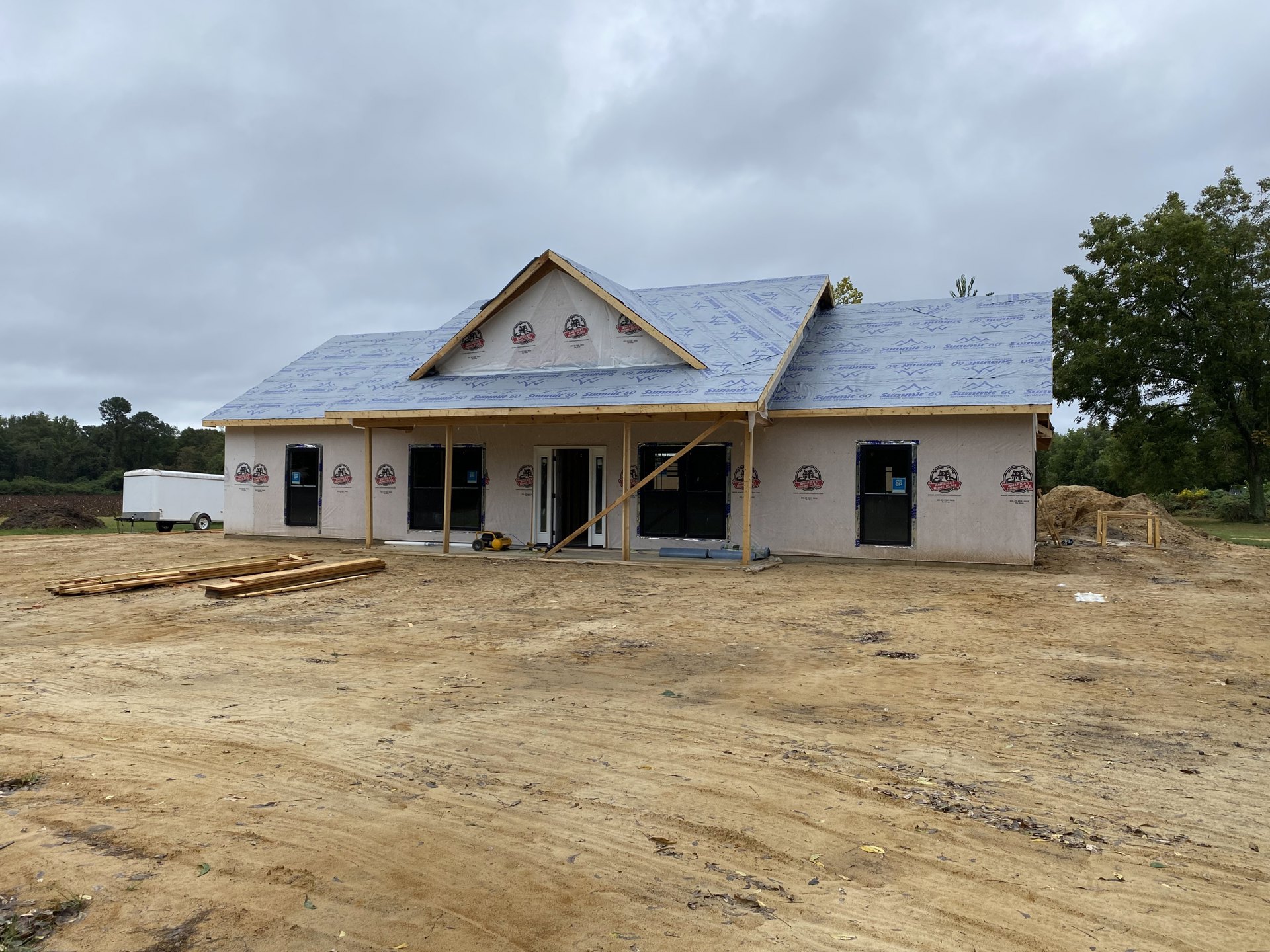 Partially built house with exposed wooden framing, triangular roof structure, dirt construction site, leafy trees in background, white trailer parked on grassy area