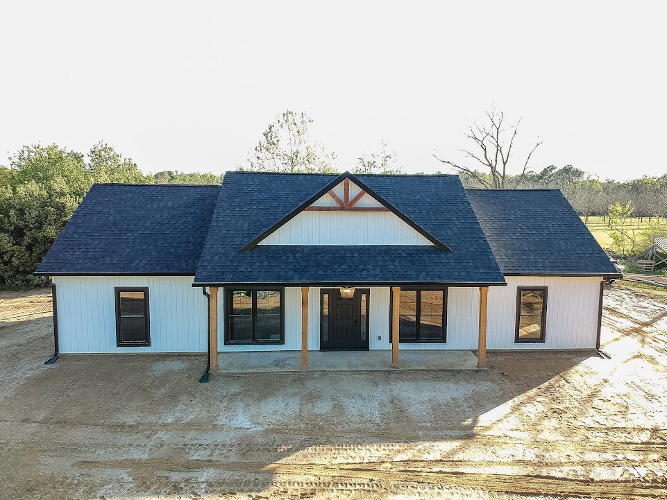 Modern home exterior with black shingle roof, black front door featuring glass panes illuminated from inside, white walls, metal-barred window, and landscaped yard with trees.