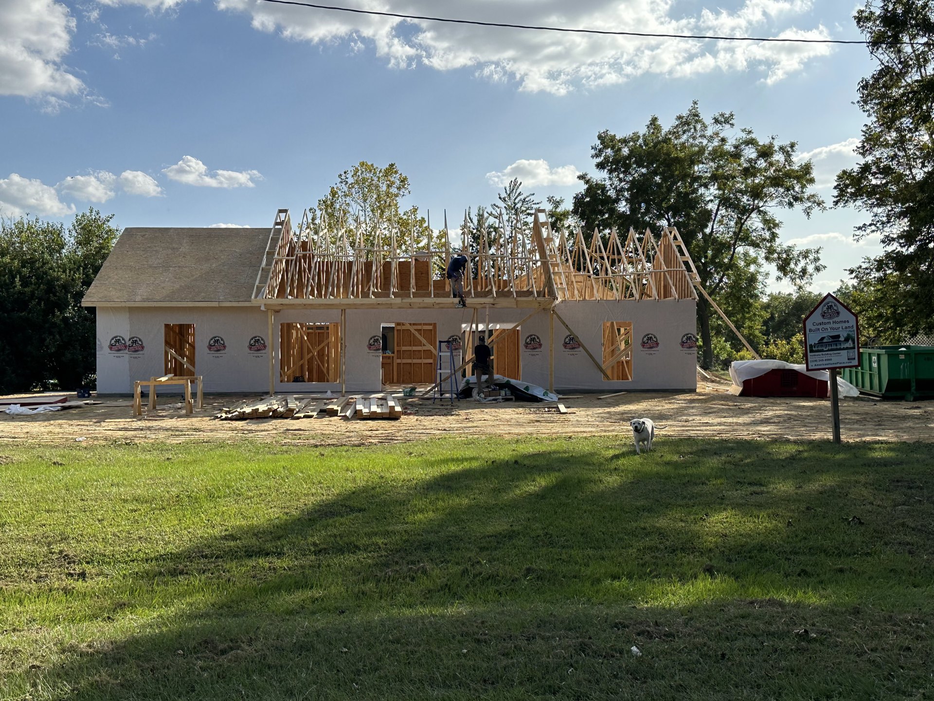 Partially built house with exposed framing, workers on the roof, grassy yard, mature trees, construction sign displaying house rendering, dog standing in lawn under cloudy sky