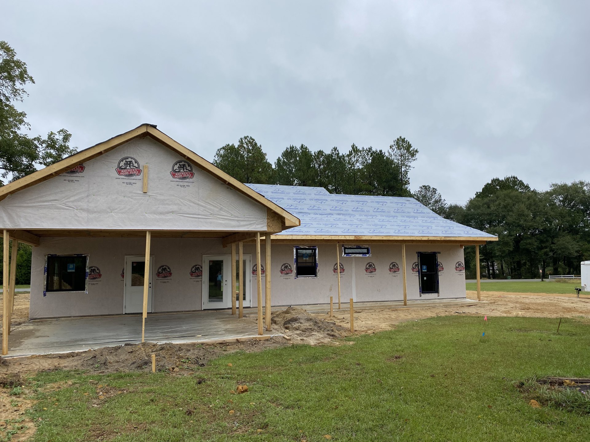 Framed house under construction with exposed wood, taped windows, dirt piles, and grassy lot bordered by trees