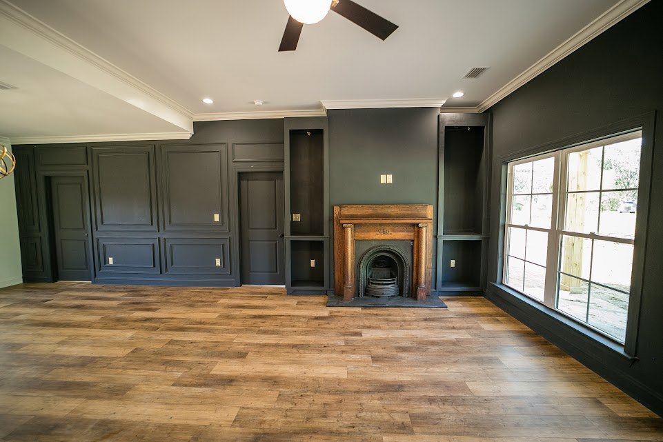 Stone fireplace with wood mantel, wide plank hardwood flooring, white walls with crown molding, large window letting in natural light, recessed ceiling lights, and a light switch