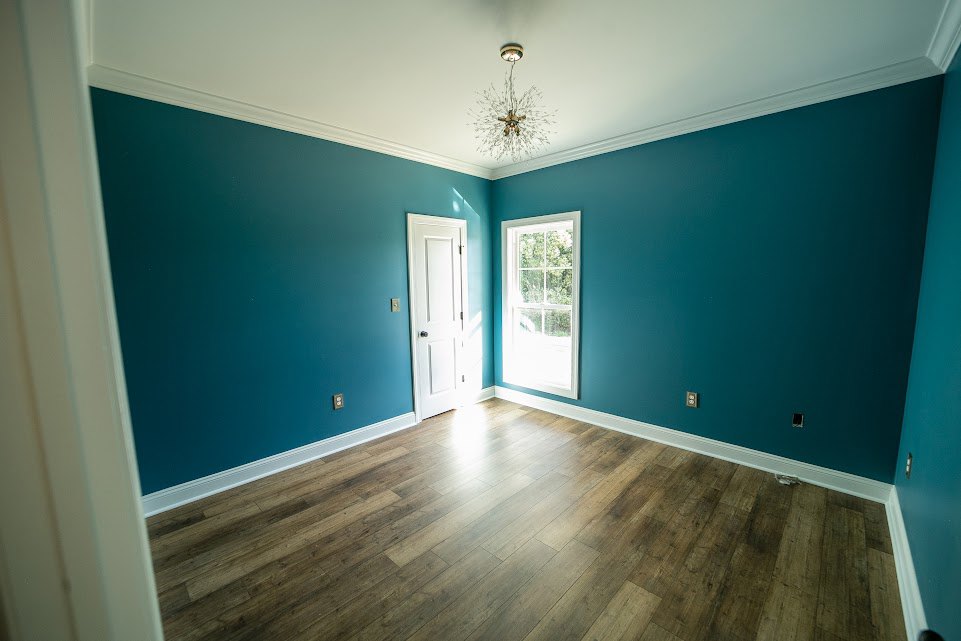Blue-painted walls, wood plank flooring, window with blue frame, white door with black knob, ceiling-mounted light fixture, white baseboard molding
