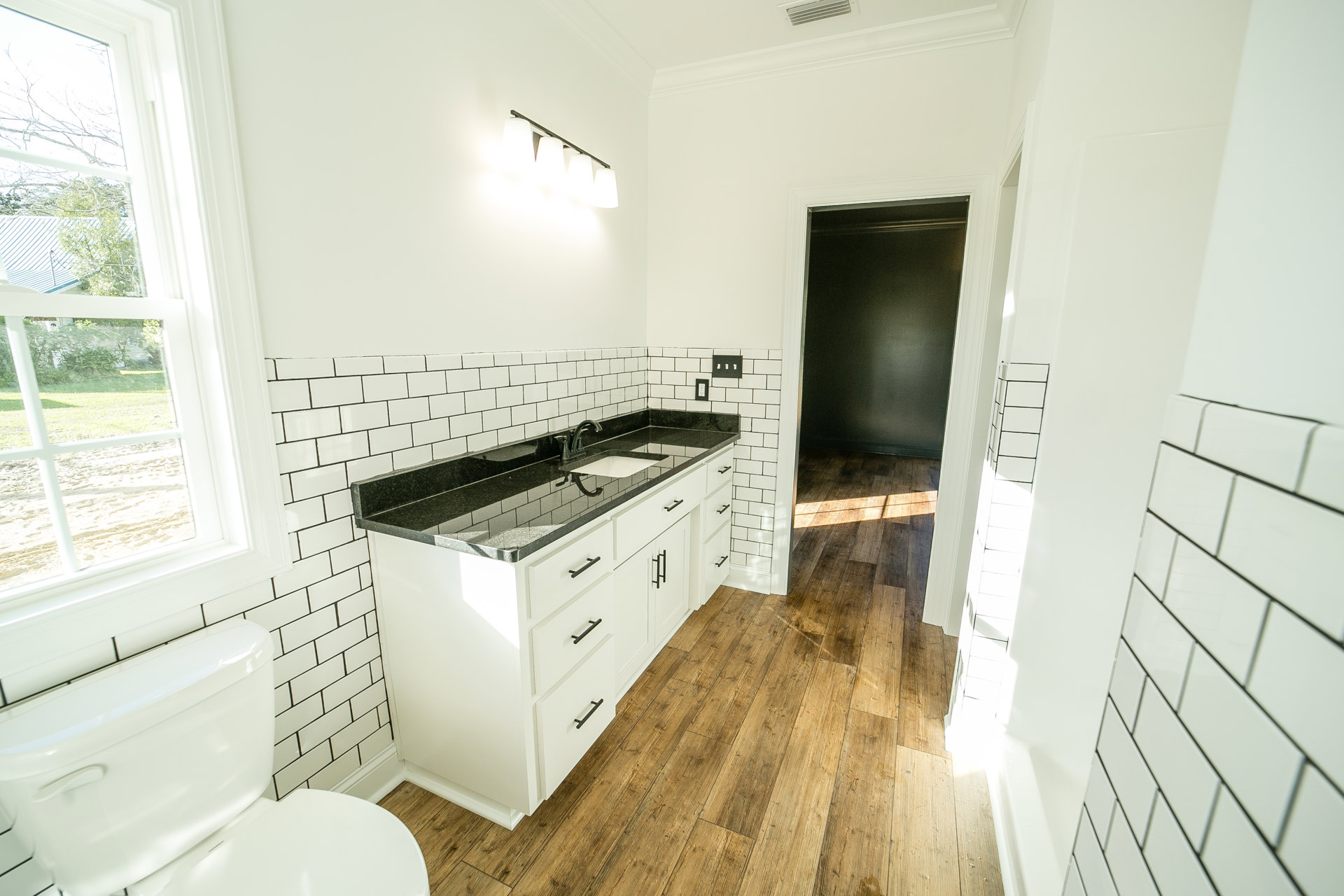 White bathroom with black granite countertop, undermount sink, white toilet, white tile walls, and window overlooking house and trees