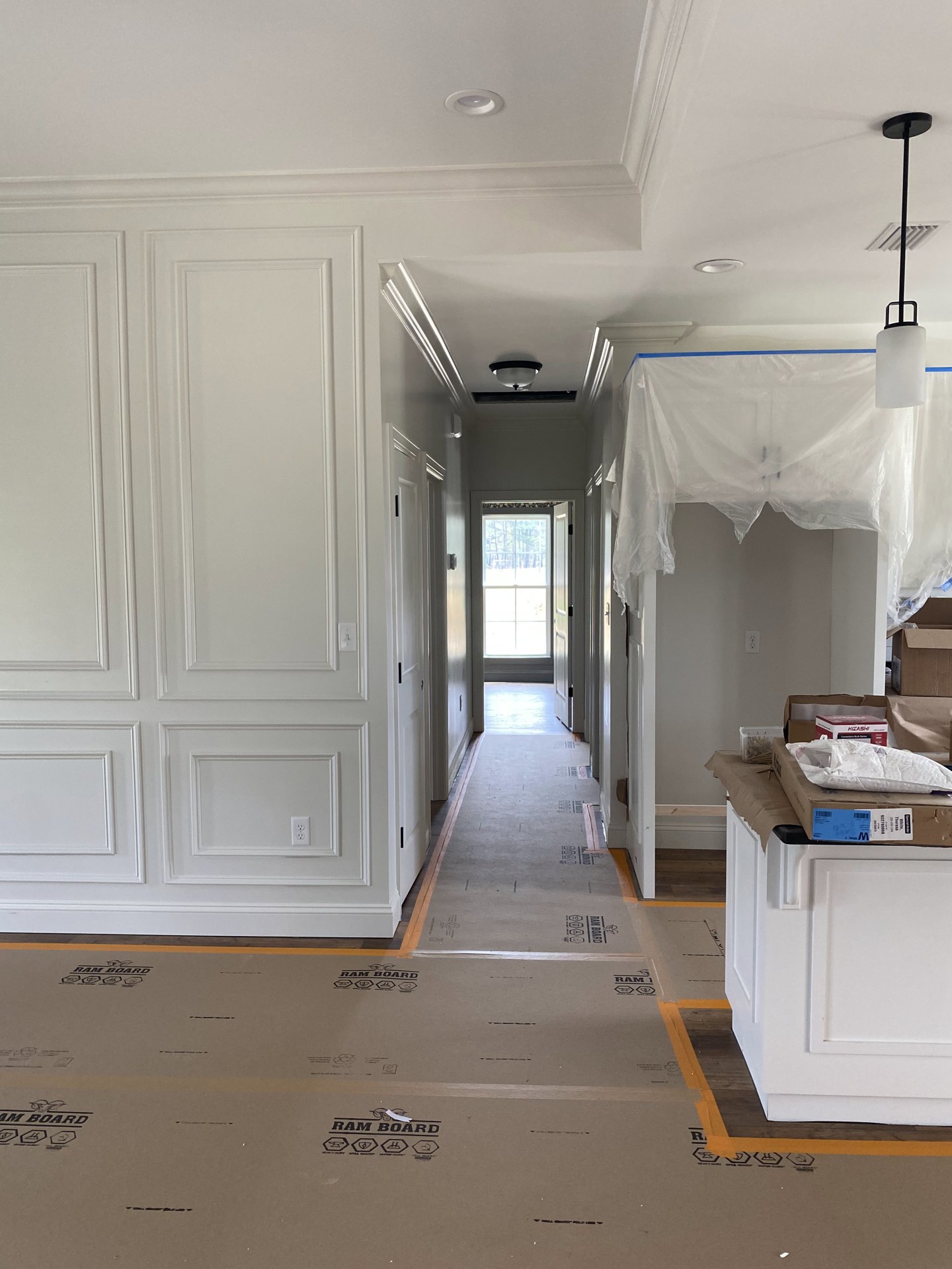 Hallway with white walls and ceiling, black pole casting a shadow, window overlooking forest, white wall with molding, plastic covering on ceiling, cardboard boxes on countertop