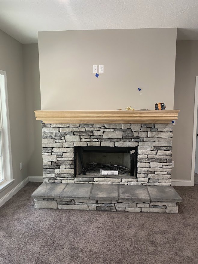 Stone fireplace with metal wood-burning stove, wood mantel shelf, and square stone hearth in a cozy den with brick wall and wood flooring.
