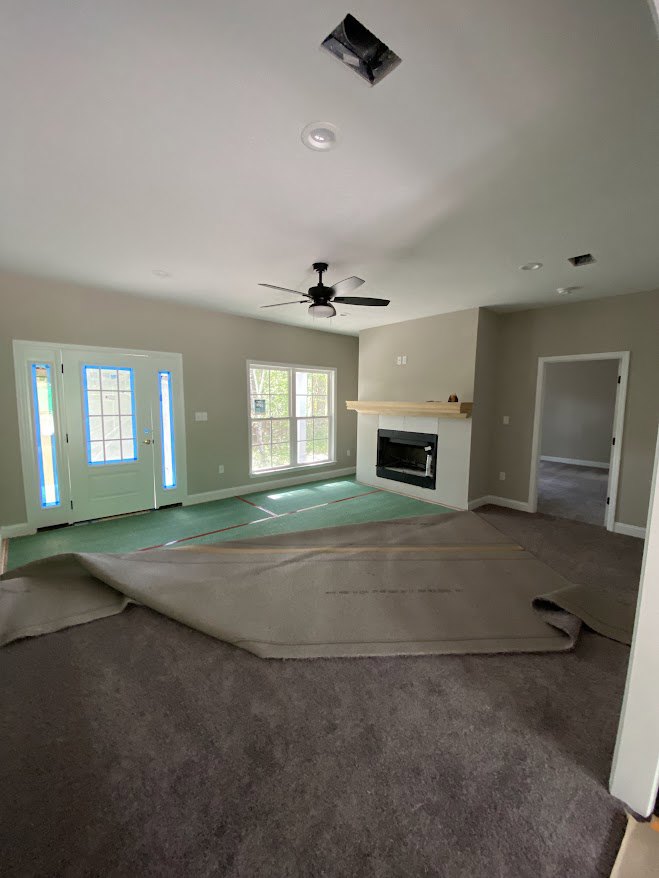 Carpeted living area with white fireplace, ceiling fan with light, multi-pane window, white door with blue trim, and open black rectangular object