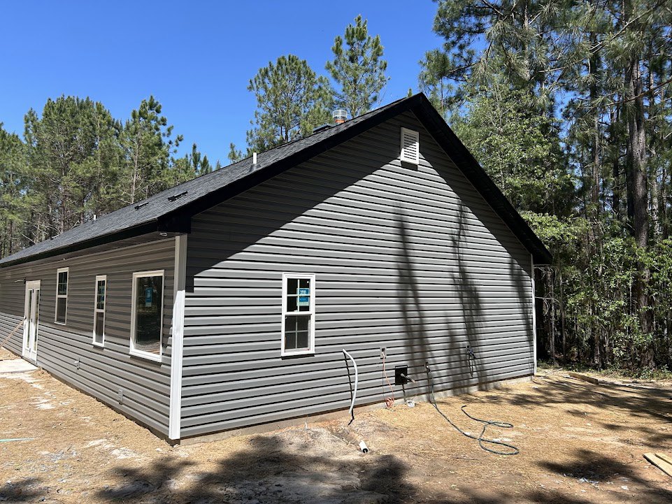 Wood-sided cottage with gabled roof, large windows, and garden hose on ground; mature trees and blue sky in background
