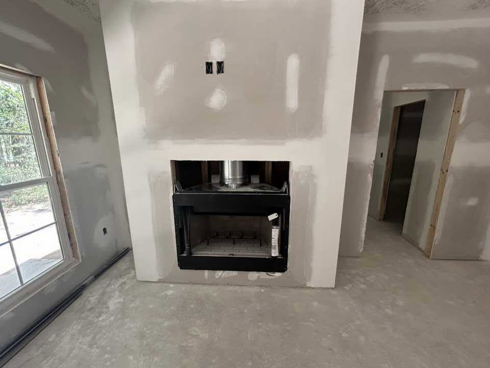 Stone fireplace with white mantel set into plaster wall, adjacent to window with white frame, hardwood floors, and neutral painted walls in living room.