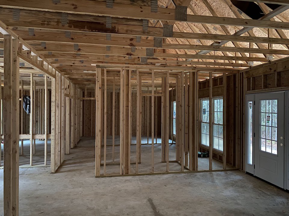 Wood-framed interior under construction with exposed beams, unfinished walls, white door and window displaying signs, and black square detail in framing